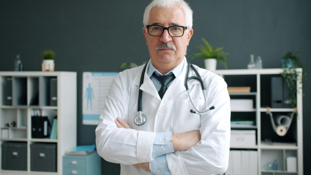 A senior doctor with white hair and a mustache wearing a lab coat and stethoscope. He stands with arms folded against a background of medical office shelving and a dark grey wall.