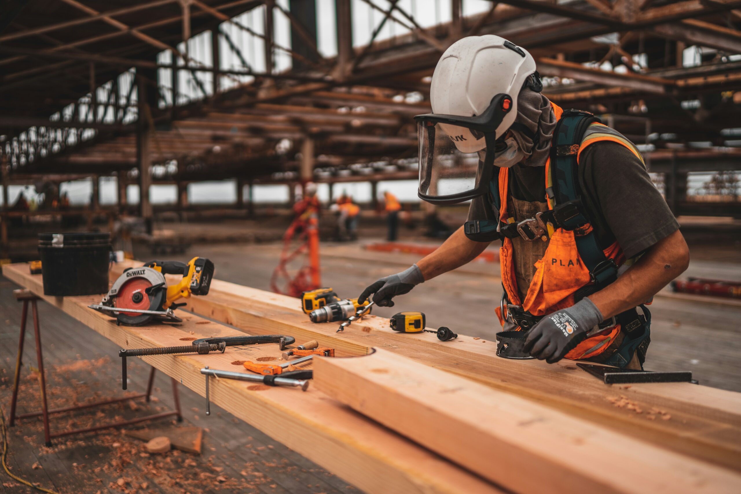 A construction worker wearing a white hard hat, face shield, and safety vest uses a drill on a large wooden beam. The workbench is scattered with tools including a circular saw, clamps, and a tape measure in an open industrial setting.