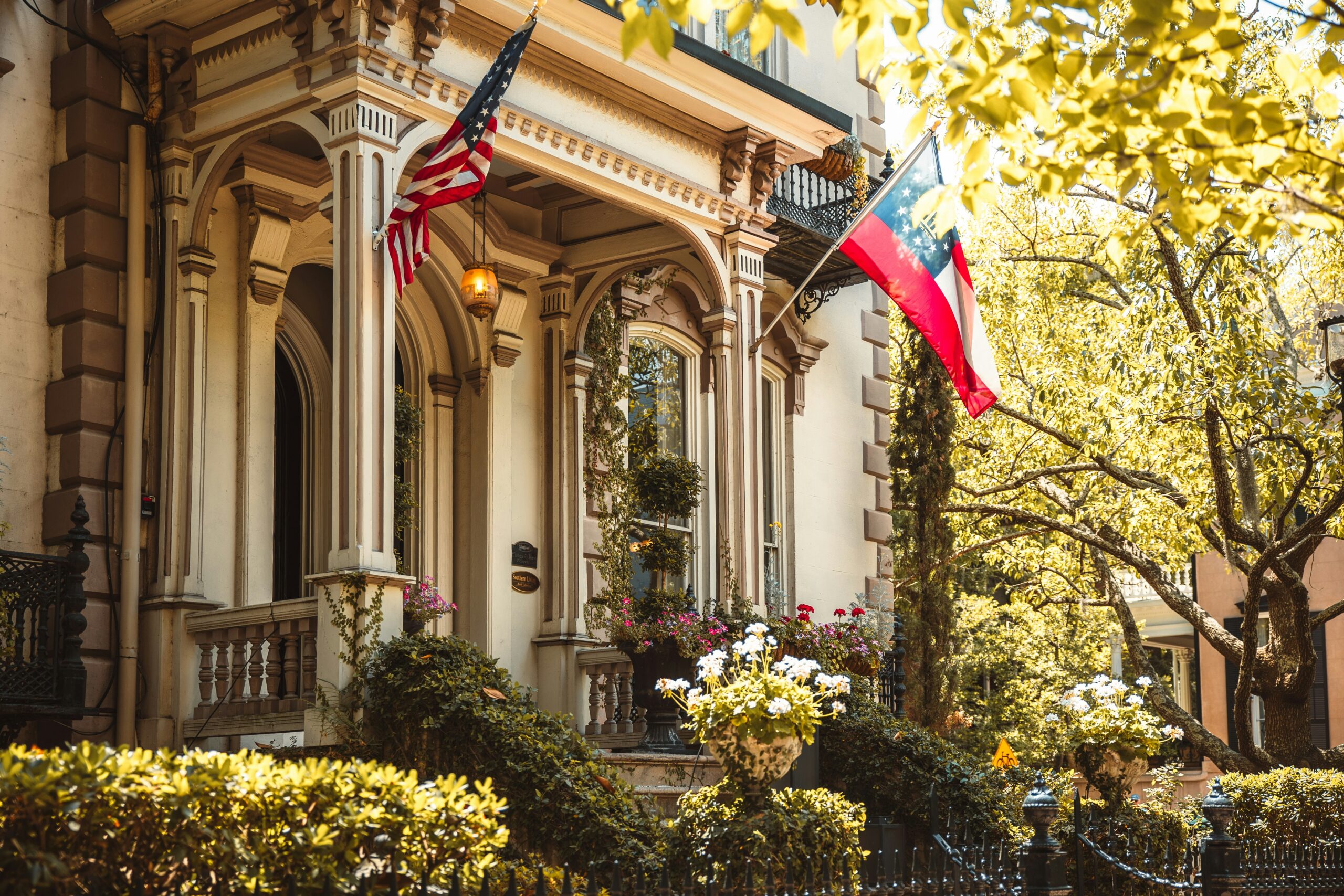 A sunlit, historic Victorian-style townhouse in Georgia featuring ornate architectural details, lush greenery, and blooming flowers. An American flag and the Georgia state flag hang prominently from the elegant front porch.