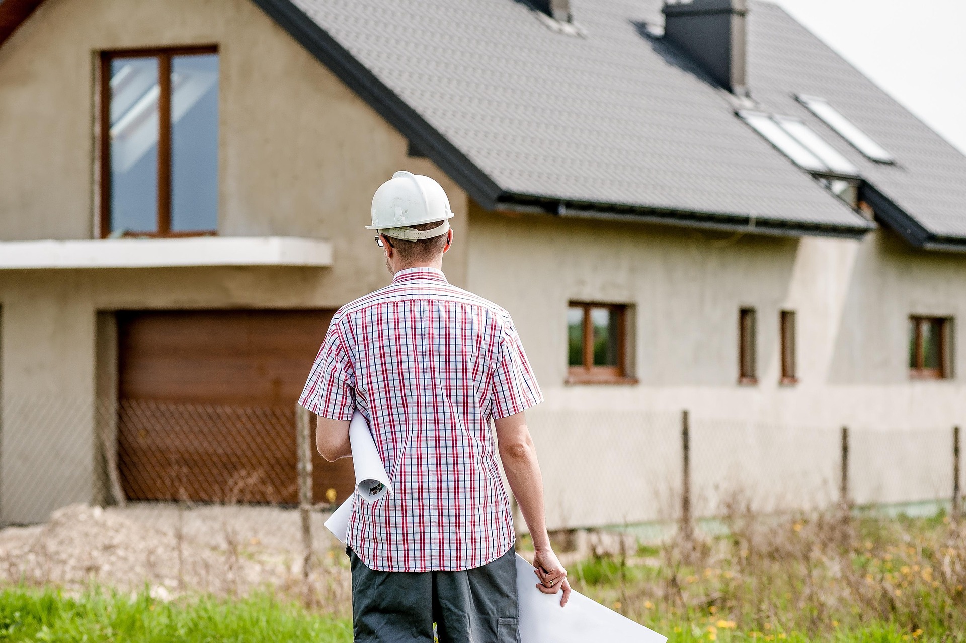 A rear view of an architect in a white hard hat and plaid shirt, carrying rolled-up blueprints while looking toward a two-story residential house under construction. The image depicts the professional responsibility and site visits that fall under liability coverage