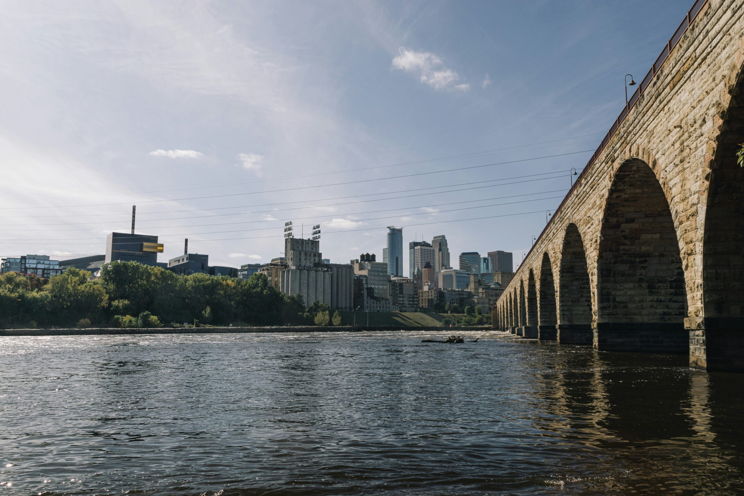 A low-angle view from the surface of the Mississippi River looking toward the Minneapolis skyline. On the right, the massive stone arches of the Stone Arch Bridge recede into the distance. In the center, the historic Gold Medal Flour grain elevator stands prominently along the riverbank, with the modern glass skyscrapers of downtown Minneapolis rising behind it. The sun shines from the left, casting a soft light over the rippling water and the city.