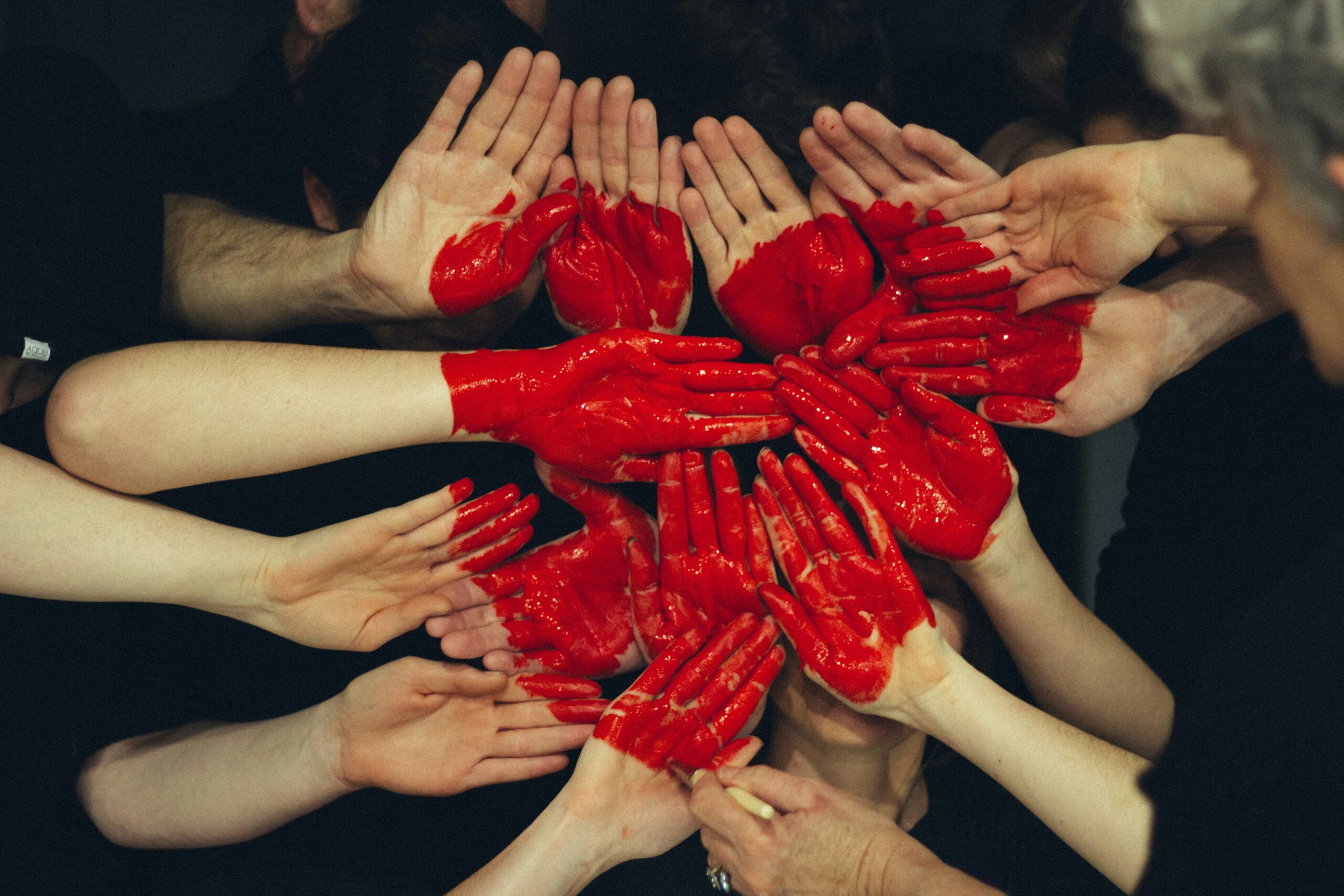 A group of people reaching their hands toward the center of the frame. Several of the palms are painted bright red, while others remain natural, creating a collaborative and artistic circular pattern against a dark background.