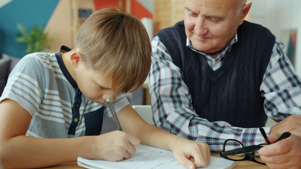 An elderly male education consultant or private tutor wearing a plaid shirt and sweater vest, providing one-on-one instruction to a young boy writing in a notebook, illustrating the need for teacher liability insurance.