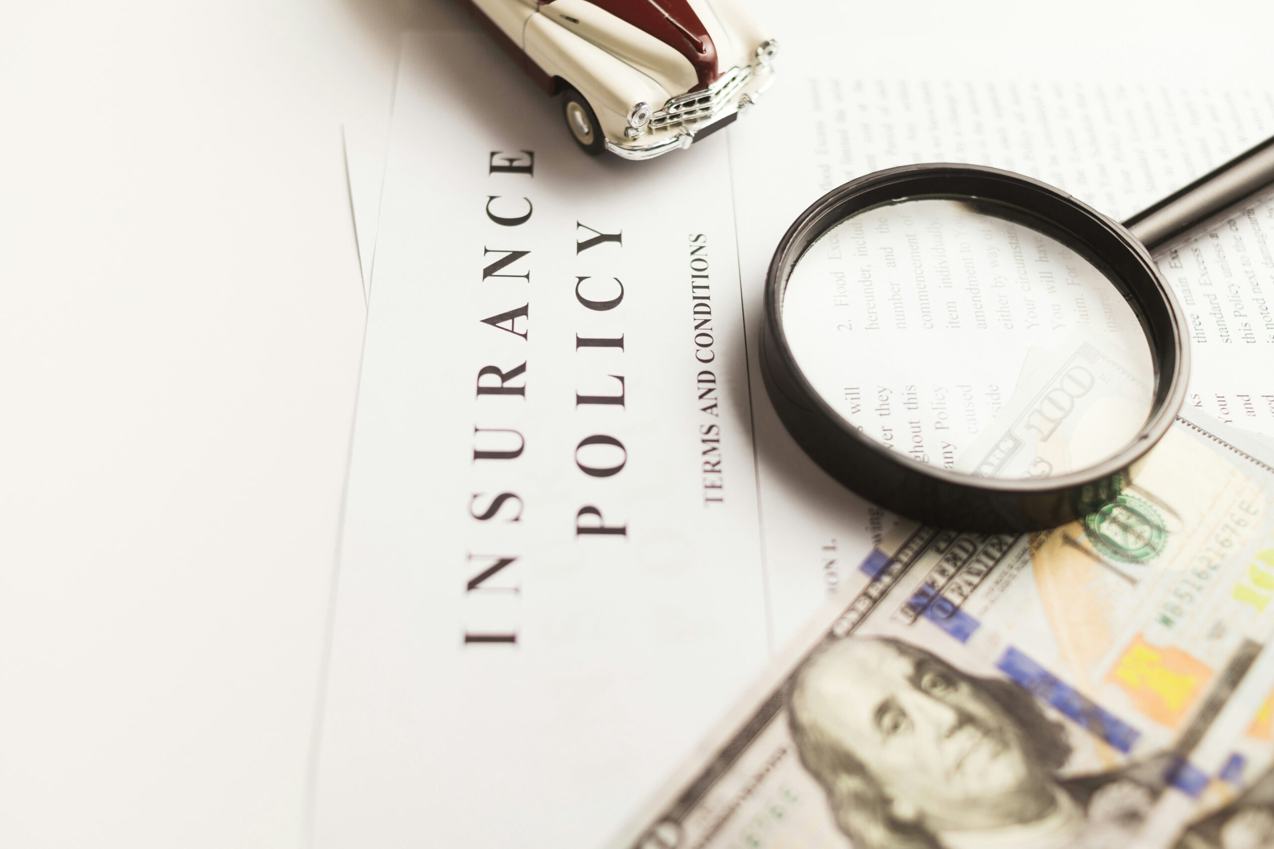 A top-down, high-angle shot of an insurance policy document labeled "INSURANCE POLICY - TERMS AND CONDITIONS." On top of the paperwork sits a small vintage model car, a magnifying glass focused on the fine print, and several one-hundred-dollar bills.