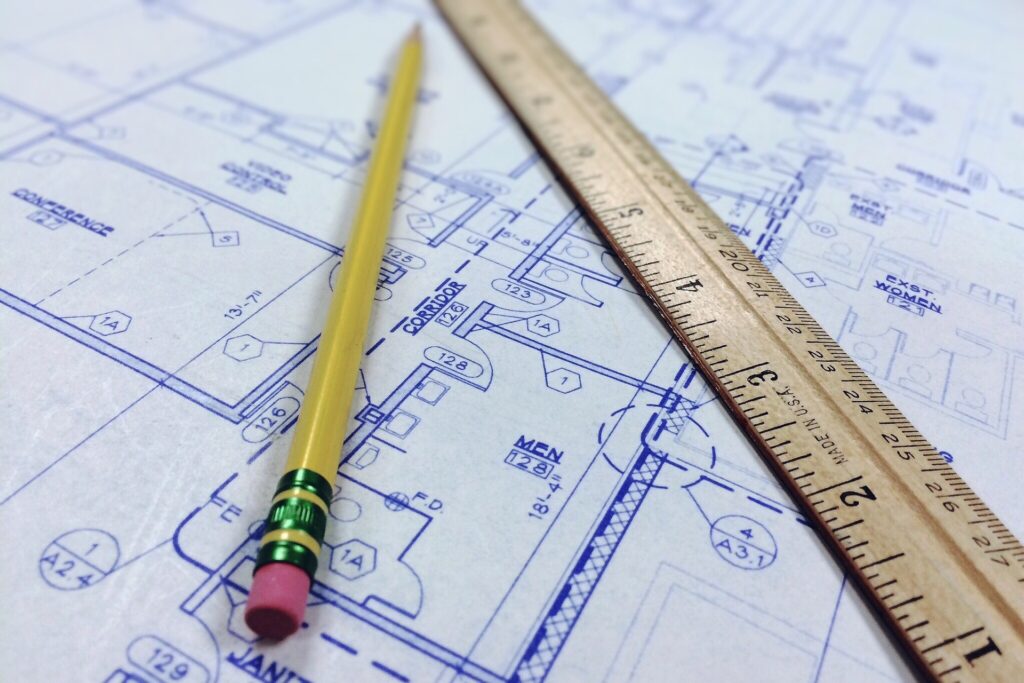A detailed close-up of a blue architectural floor plan on a desk, featuring a yellow pencil with a pink eraser and a wooden ruler. The image represents the precision and technical documentation required in architectural design that professional liability insurance protects.